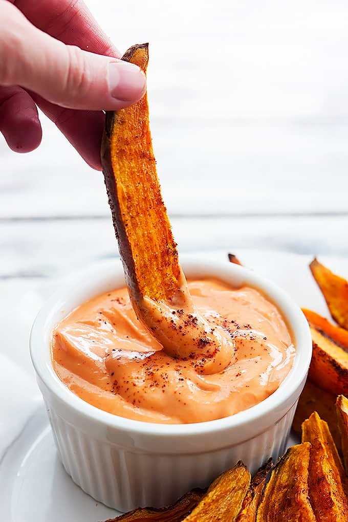 a hand dipping a spicy baked sweet potato wedge in a dipping bowl of fry sauce.