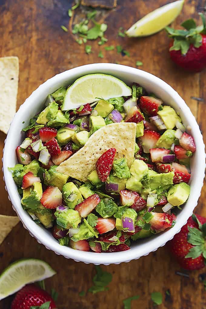 top view of strawberry avocado salsa in a bowl with a chip dipped inside surrounded by chips, lime slices and strawberries.