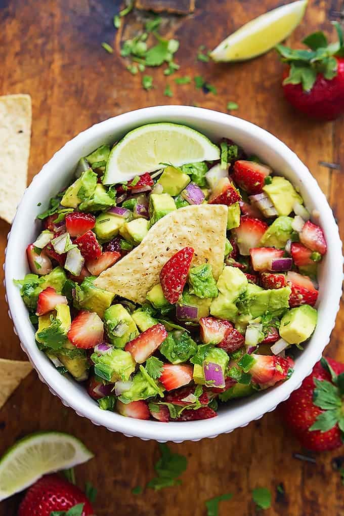 top view of strawberry avocado salsa in a bowl with a chip dipped inside surrounded by chips, lime slices and strawberries.