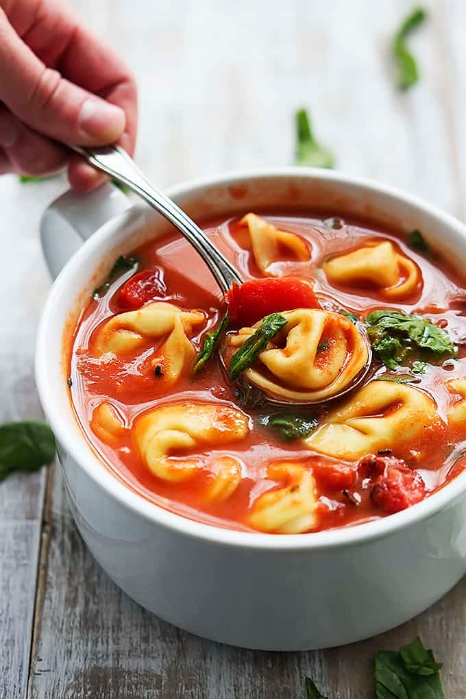 a hand scooping some tomato spinach tortellini soup out of a bowl with a spoon.