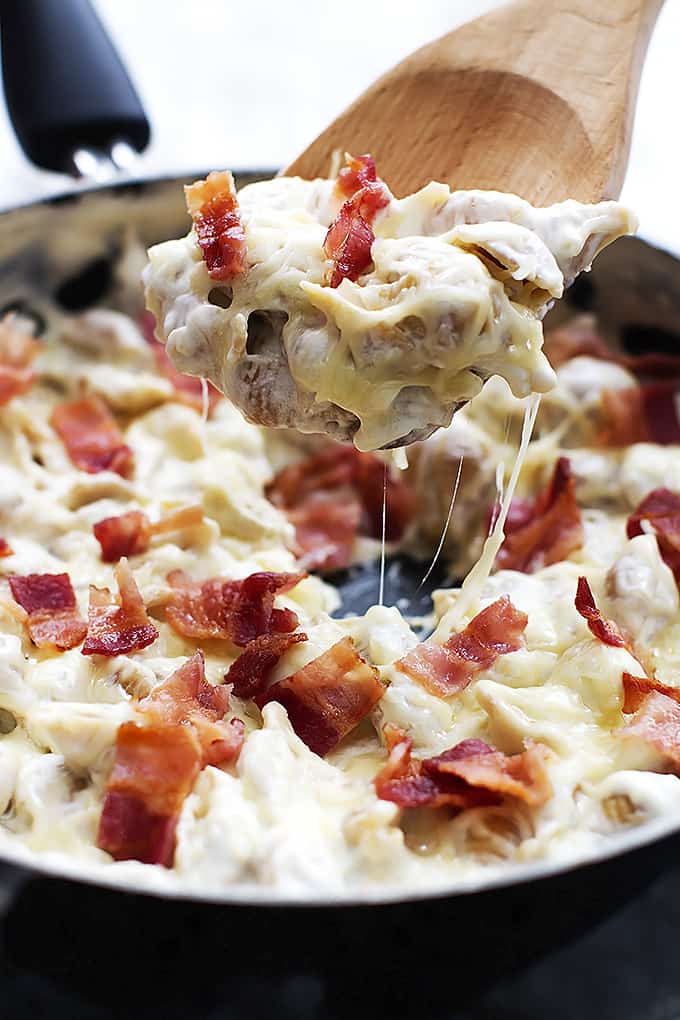 Close up of a wooden serving spoon with a scoop of cheesy bacon alfredo shells above a skillet full of more shells.