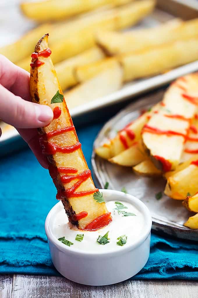 a hand dipping a baked garlic sriracha fry in dipping sauce with more fries in the background.