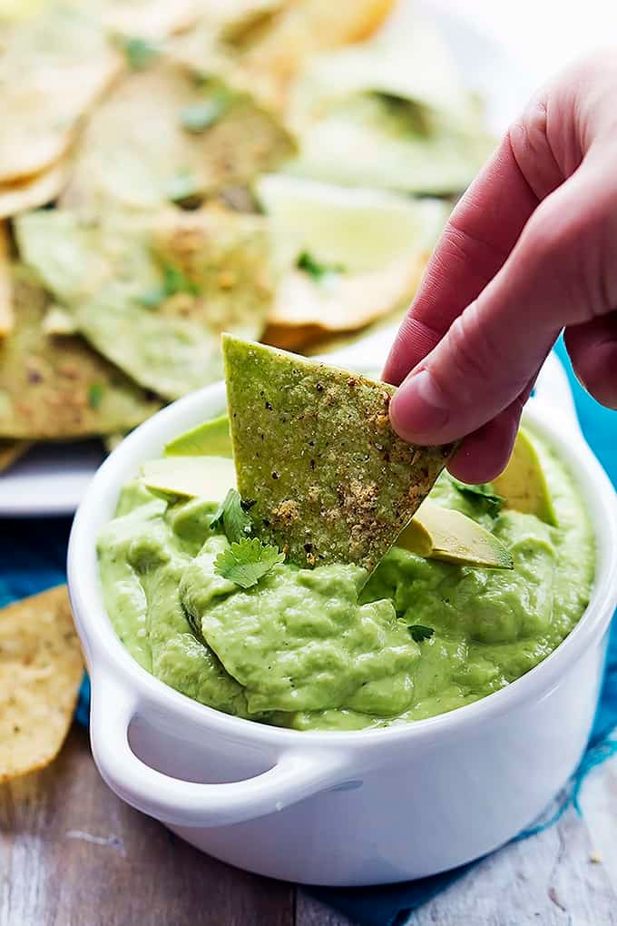 a hand dipping a baked guacamole tortilla chip in a bowl of guacamole with a plate of more chips in the background.