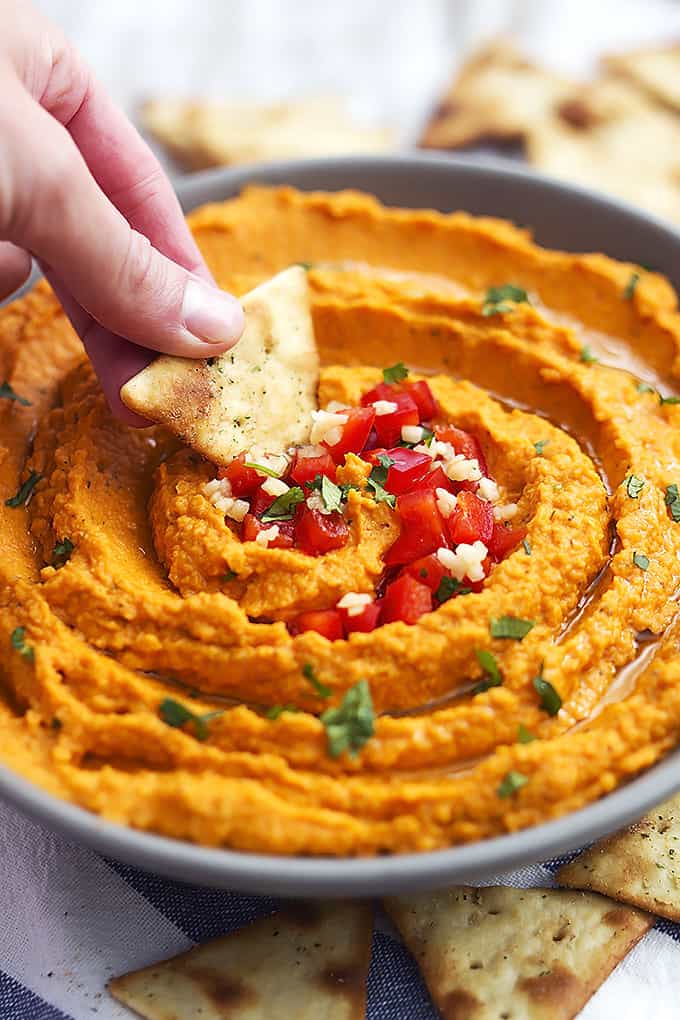 a hand dipping a chip in roasted red pepper hummus in a bowl.