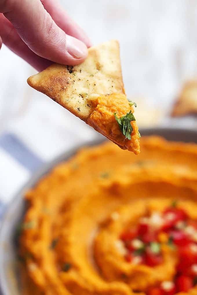 a hand holding a chip dipped in roasted red pepper hummus above a bowl of hummus.