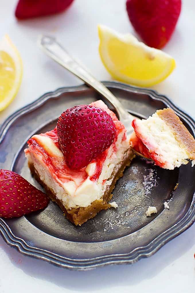a strawberry lemon cheesecake bar with a bite on a fork on the side all on a plate with strawberries and lemon slices in the background.