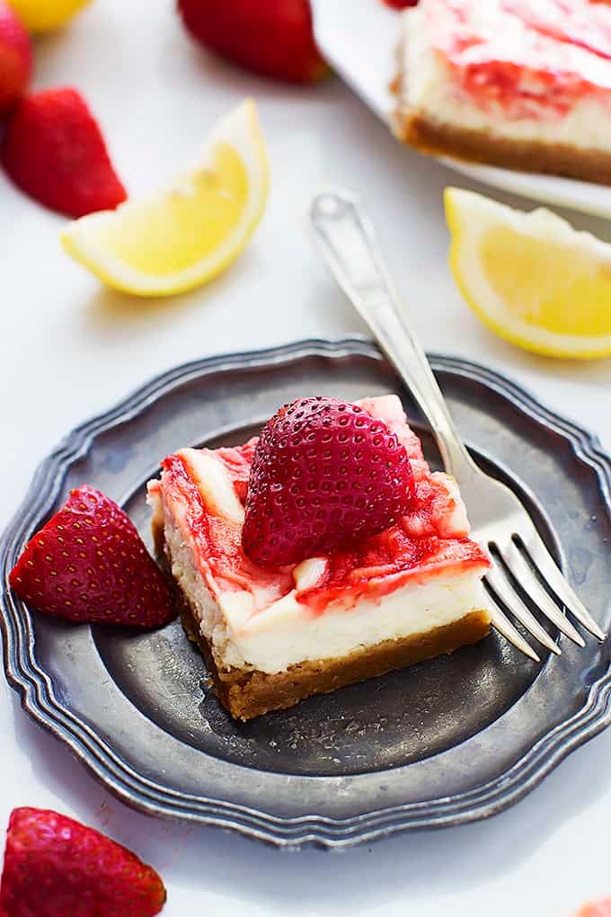 a strawberry lemon cheesecake bar with a fork on a plate with strawberries and lemon slices in the background.