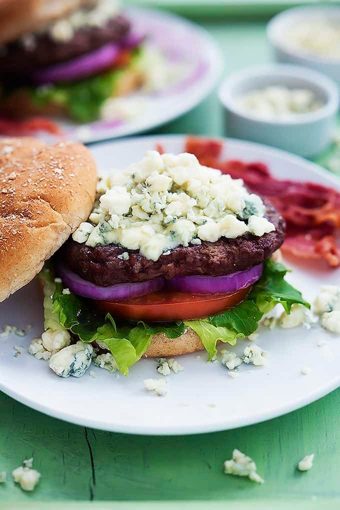 a bacon blue cheese burger on a plate with the top bun off and another burger on a plate with blue cheese in small bowls in the background.