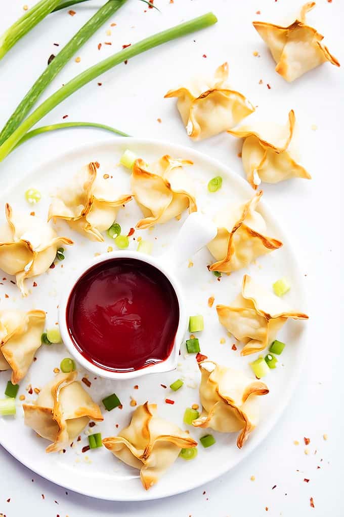 top view of baked cream cheese wontons on a plate surrounding a bowl of sauce with more wontons on the side.