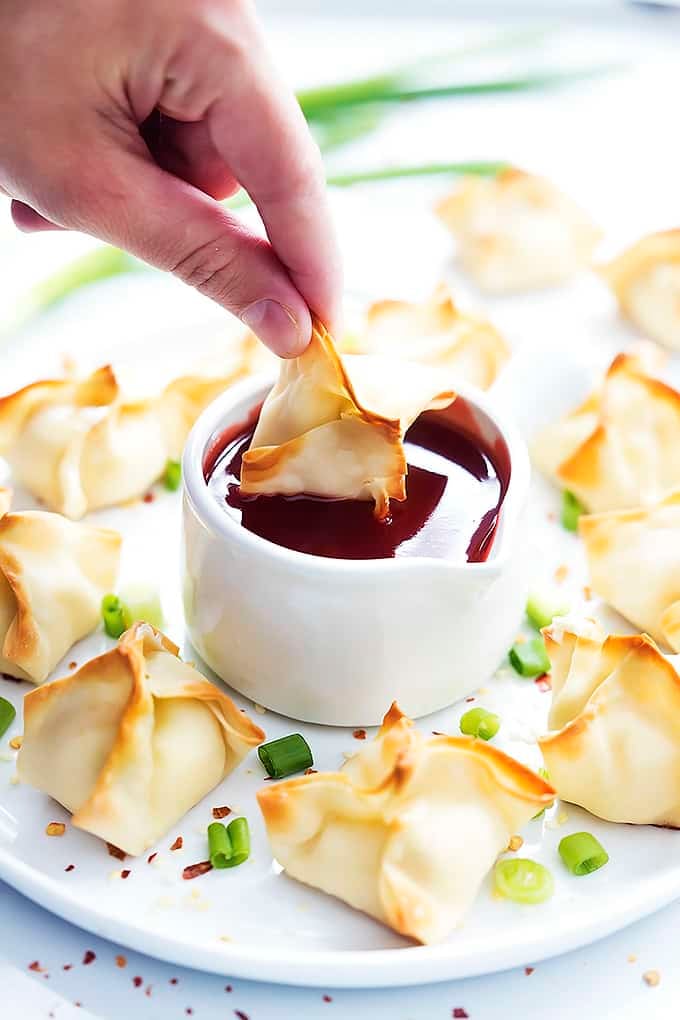 a hand dipping a baked cream cheese wonton in sauce with more wontons around it on a plate.