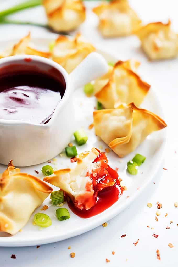 close up of baked cream cheese wontons surrounding a bowl of suace on a plate with the from wonton being dipped in sauce with a bite missing.