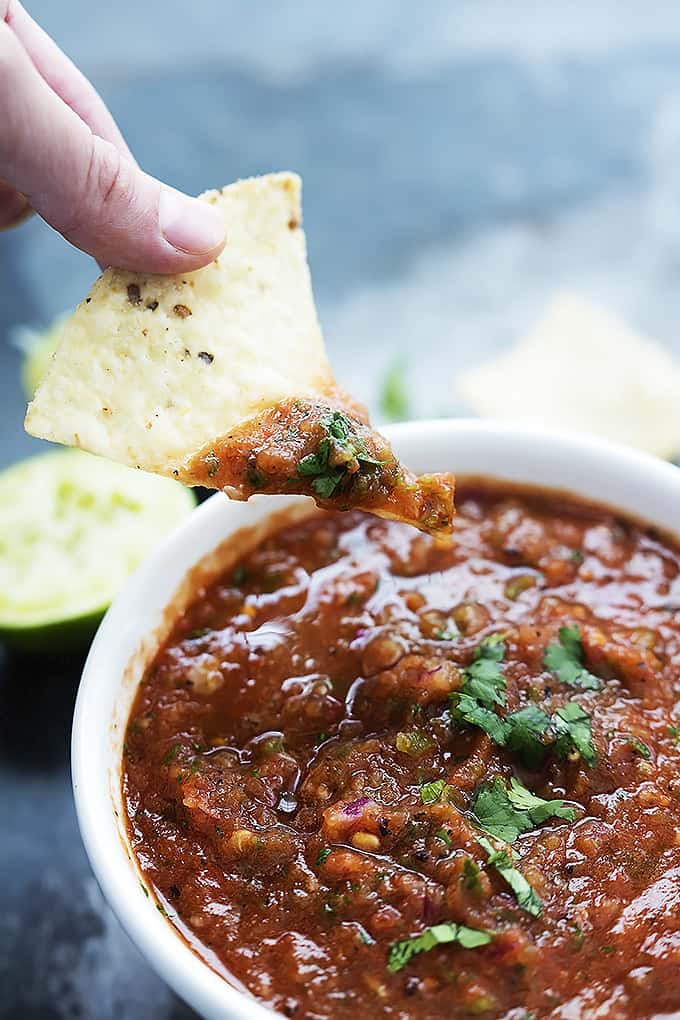 a hand holding up a chip dipped in fire roasted salsa above a bowl of more salsa with a half of a lime in the background.