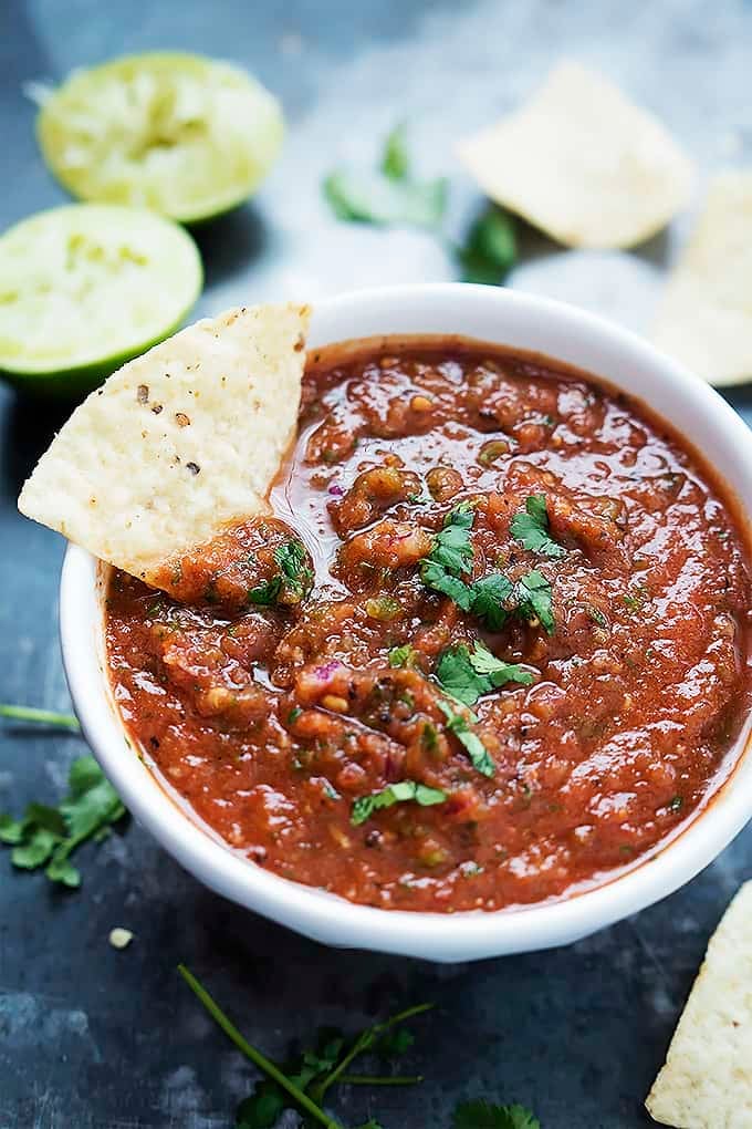 fire roasted salsa in a bowl with a chip dipped inside with two squeezed halves of lime with more chips in the background.