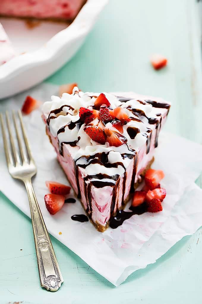 frozen strawberry cheesecake topped with cream, chocolate syrup and chopped strawberries with a fork on the side.
