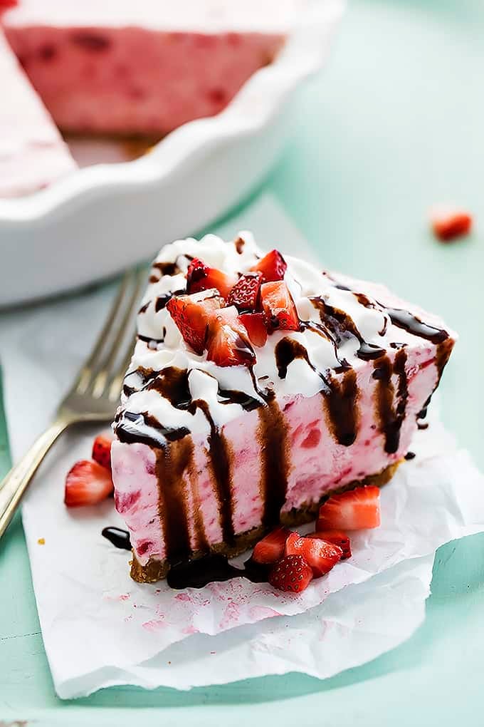 a slice of frozen strawberry cheesecake topped with cream, chocolate sauce and chopped strawberries with a fork and the rest of the cheesecake in a pit tin in the background.