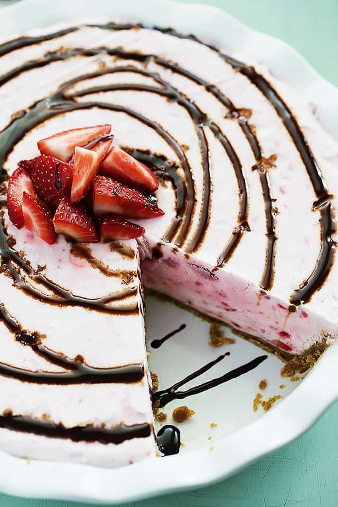 top view of frozen strawberry cheesecake in a pie tin missing a slice topped with chocolate syrup and chopped strawberries.