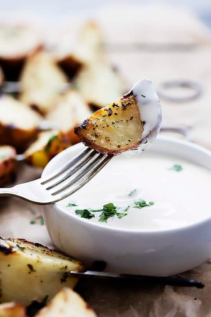 a grilled ranch potato on a fork dipped in ranch being held above a bowl of ranch with more potatoes in the background.