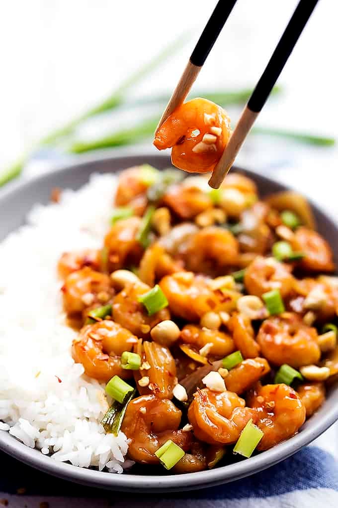 a piece of kung pao shrimp being held up by chopsticks above a plate of rice and more shrimp.