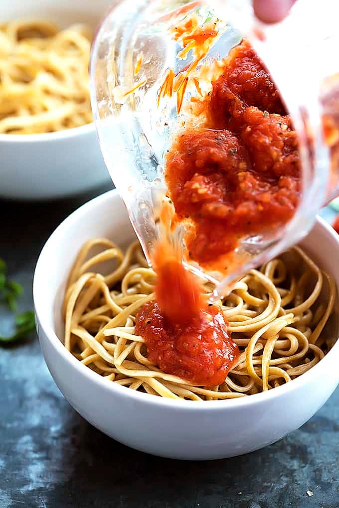 a hand pouring fire roasted tomato pasta sauce over pasta in a bowl.