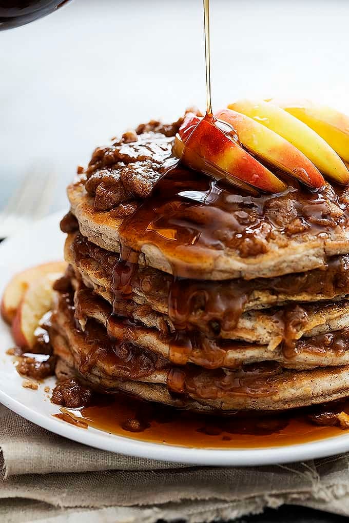 close up of a stack of apple cinnamon streusel pancakes on a plate with buttery cinnamon streusel topping and apples with syrup being poured on top.