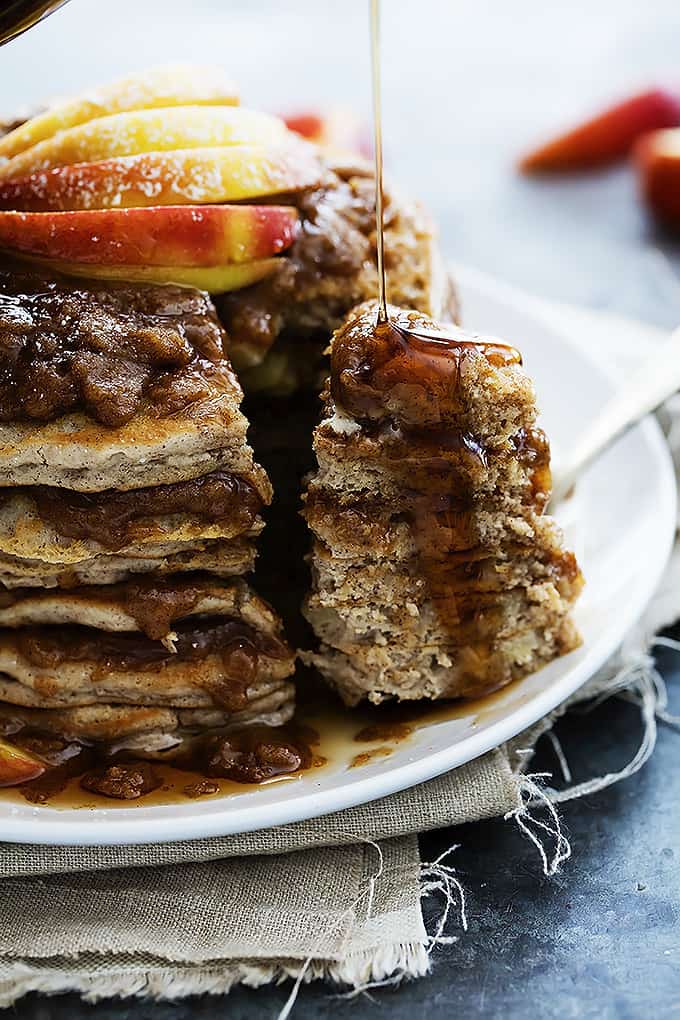 close up of a stack of apple cinnamon streusel pancakes on a plate with a chunk of pancakes separated from the rest with syrup being poured on top.