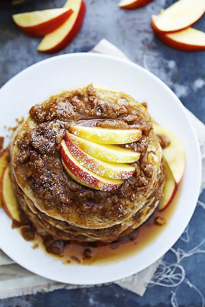 top view of a stack of apple cinnamon streusel pancakes on a plate with buttery cinnamon streusel topping, apples and syrup with more apple slices on the side.