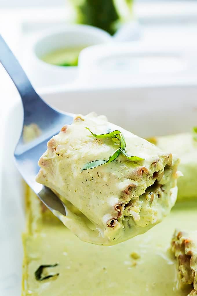 close up of a spatula holding a creamy pesto chicken lasagna roll above a cooking tray of more lasagna rolls.