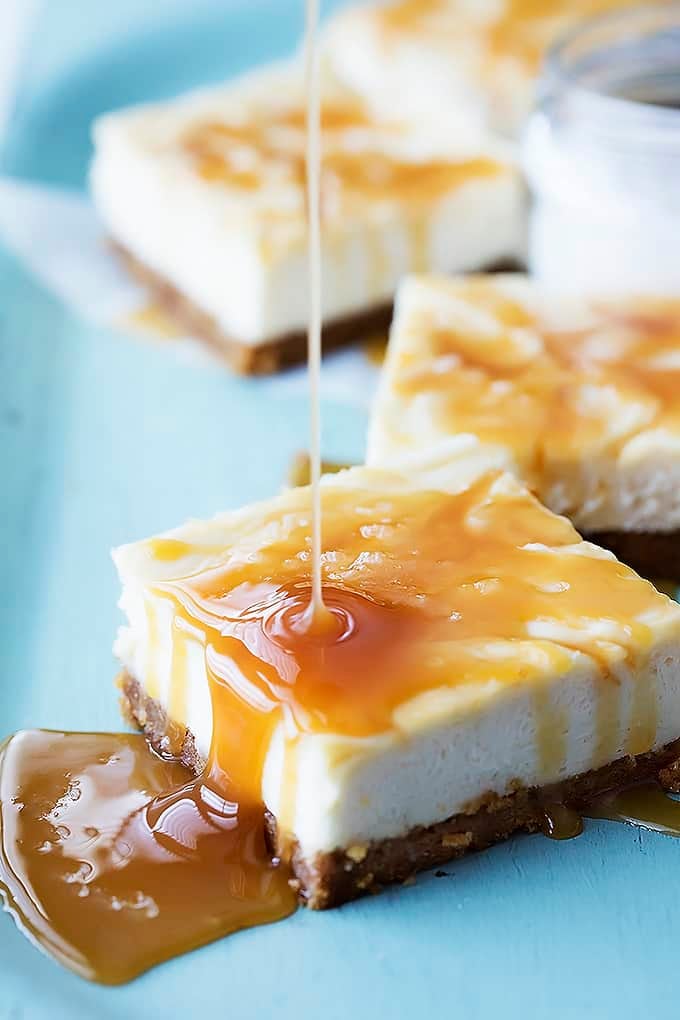 a salted caramel cheesecake bar with caramel being poured on top with more bars in the background.
