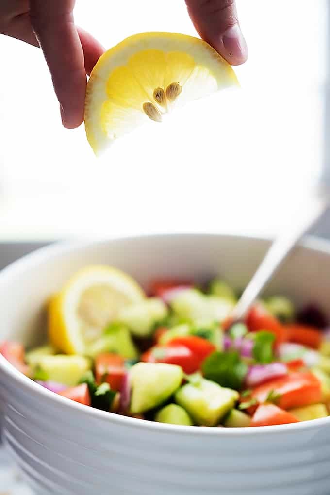 a hand holding a lemon slice above a bowl of tomato cucumber salad with a spoon.