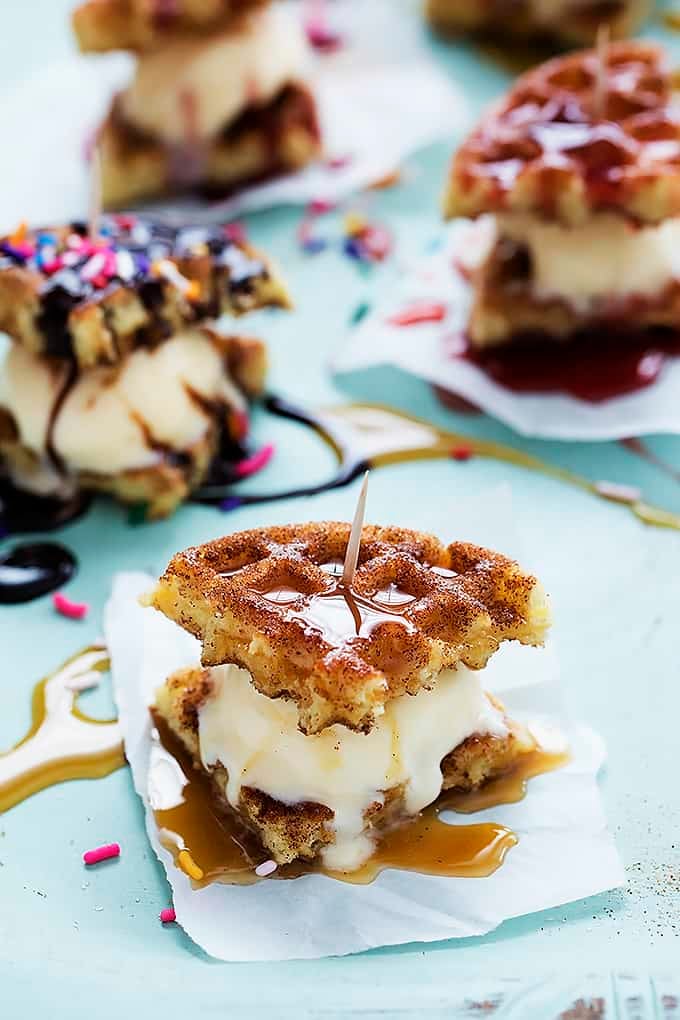 a snickerdoodle waffle ice cream sandwich with more sandwiches in the background with one being topped with chocolate syrup and sprinkles instead of caramel sauce.