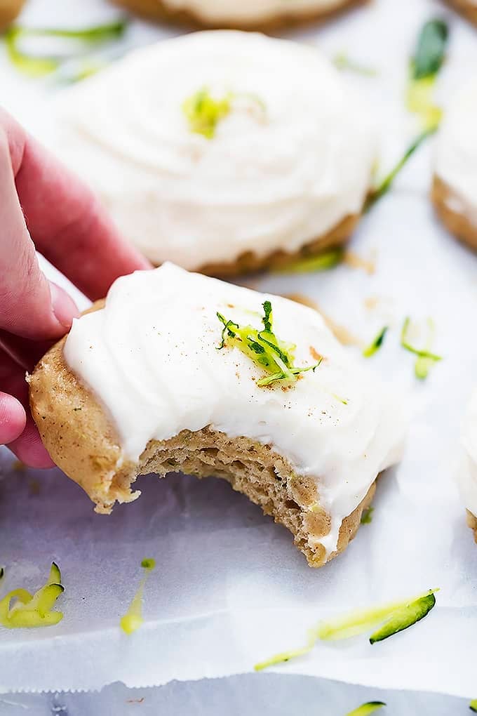 a hand putting a zucchini cookie with brown butter cream cheese frosting down with a bite missing with more cookies in the background.