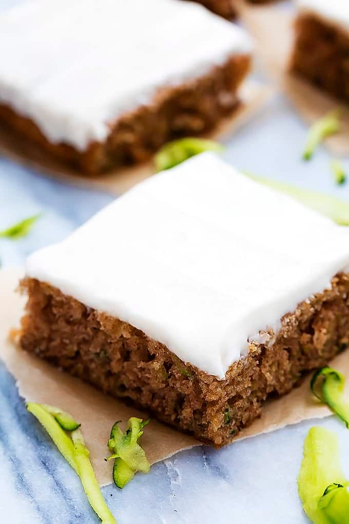 a piece of zucchini sheet cake with more pieces in the background with zucchini shavings around them.