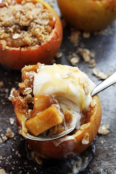 an apple crisp stuffed baked apple topped with ice cream and a spoon with a bite on it with more stuffed apples in the background.
