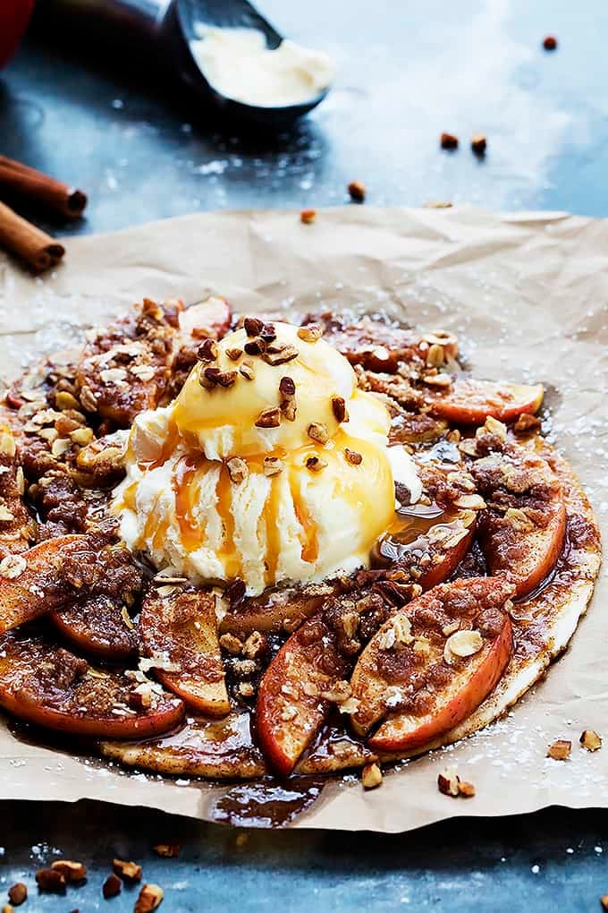 apple crumble flatbread topped with vanilla ice cream, caramel sauce and nuts with cinnamon sticks and a scoop of ice cream on an ice cream scooper in the background.