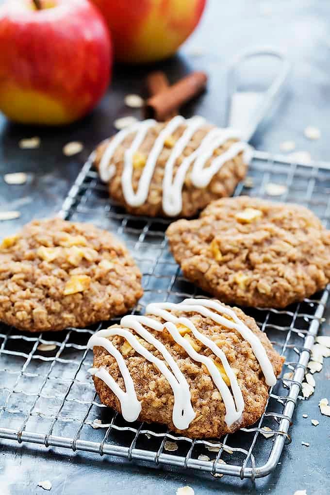 apple oatmeal cookies with some drizzled with apple cream cheese frosting on a cooling rack with whole apples, oats, and cinnamon sticks on the side.