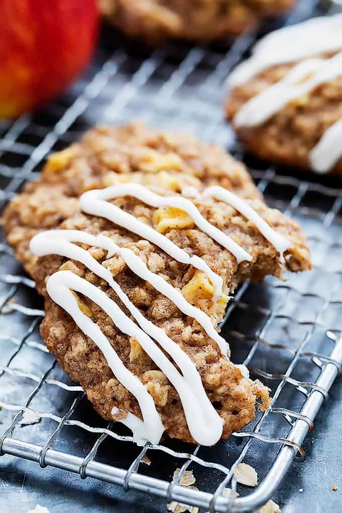 a apple oatmeal cookie drizzled with apple cream cheese frosting with a bite missing leaning on top of another cookie on a cooling rack.