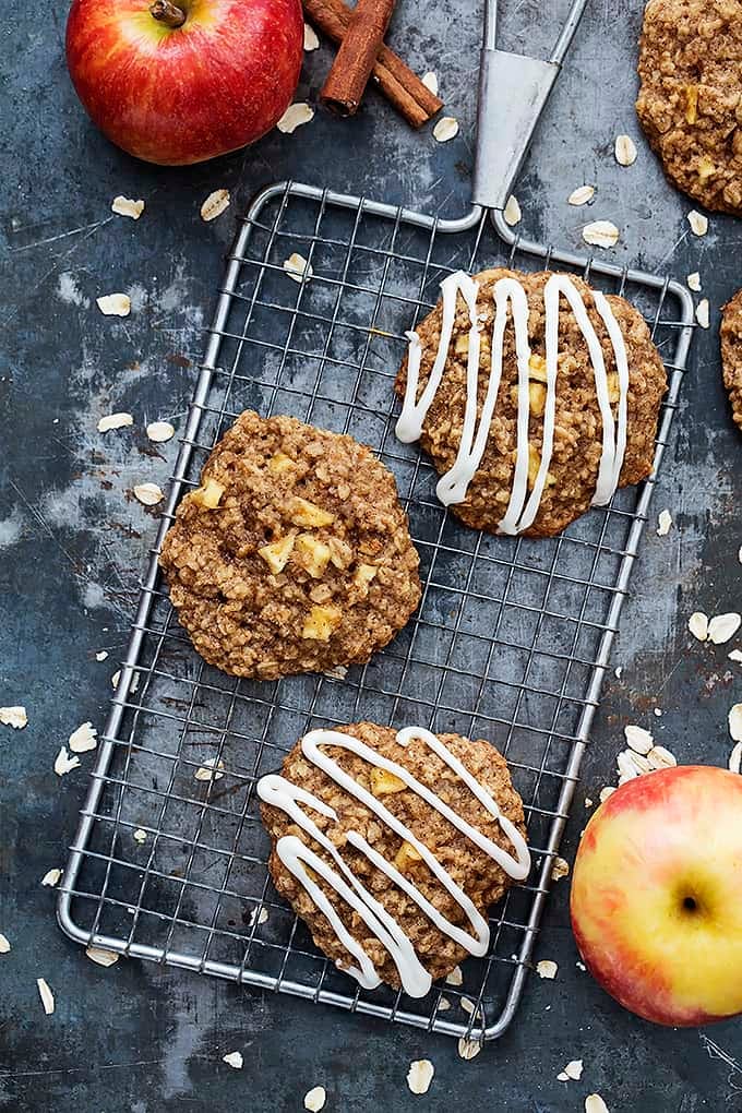 top view of apple oatmeal cookies with some drizzled with apple cream cheese frosting with more cookies, oats, apples, and cinnamon sticks on the side.