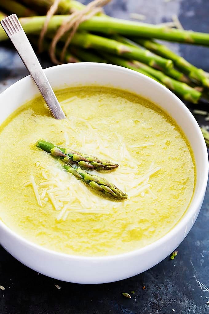 asparagus parmesan soup and a spoon in a bowl with asparagus stalks in the background.