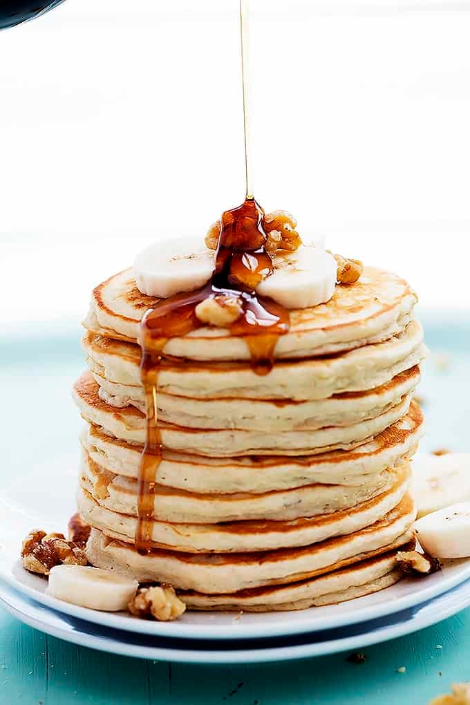 a stack of banana bread pancakes topped with bananas and walnuts with syrup being poured on top.