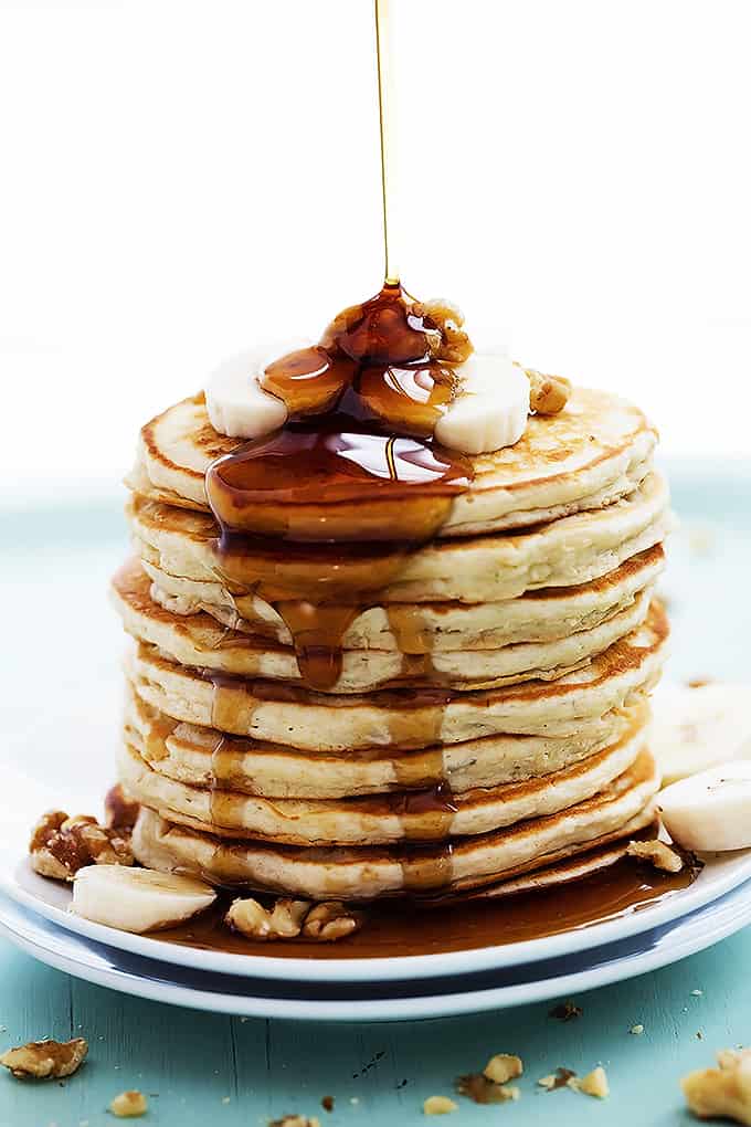 a stack of banana bread pancakes on a plate topped with bananas and walnuts with syrup being poured on top.