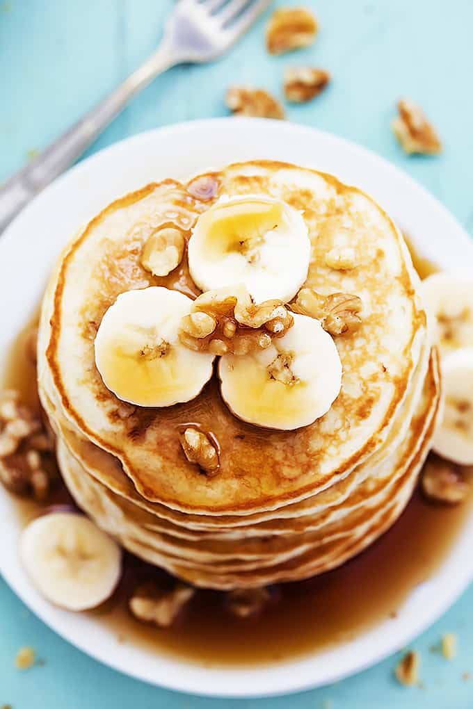 close up top view of a stack of banana bread pancakes on a plate topped with bananas, walnuts, and syrup with more walnuts and a fork on the side.