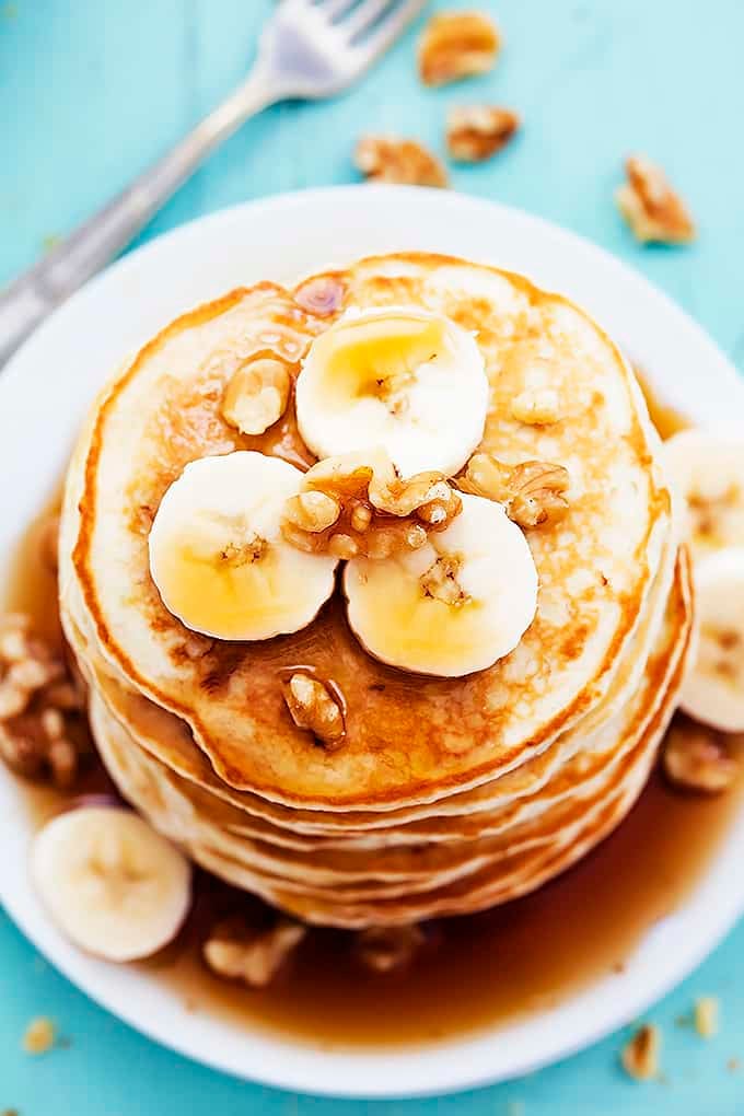 close up top view of a stack of banana bread pancakes on a plate topped with bananas, walnuts, and syrup with more walnuts and a fork on the side.