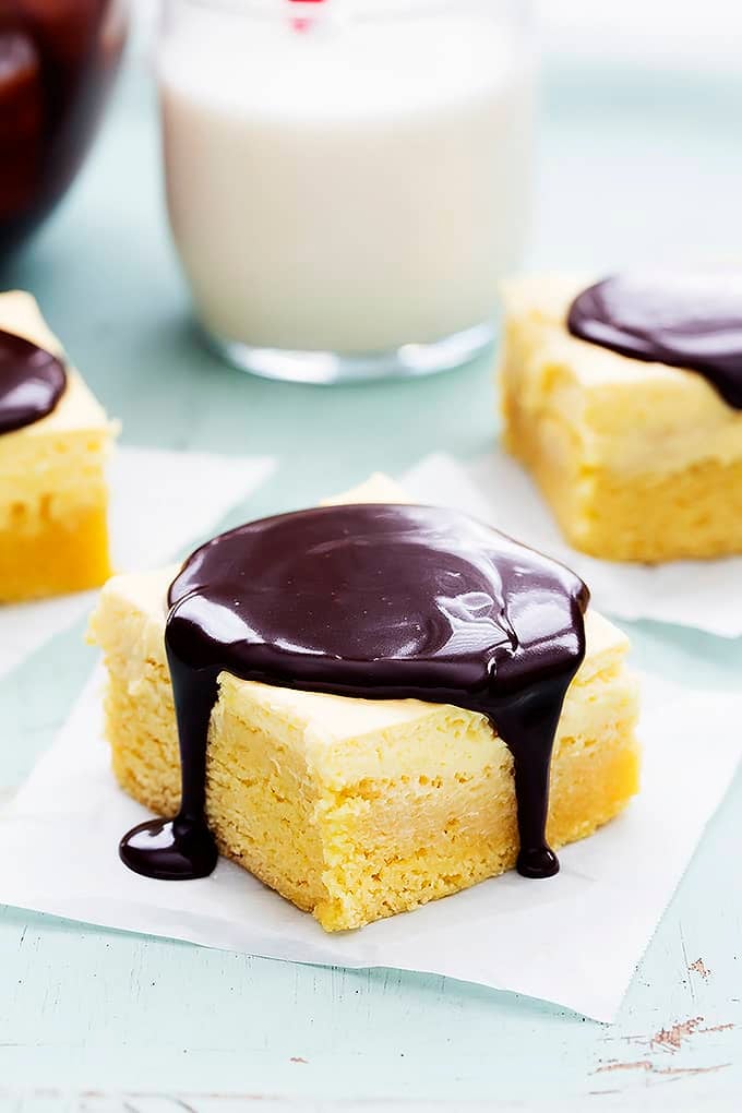 a Boston cream pie bar with chocolate ganache on top with more bars with ganache and a glass of milk in the background.