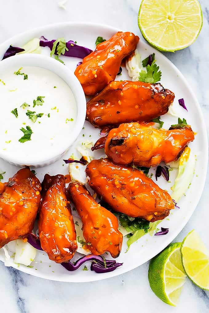 top view of spicy honey lime chicken wings next to a bowl of dipping sauce on a plate with a lime half and lime slices on the side.