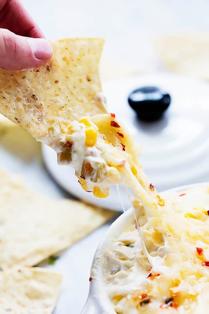 a hand pulling a chip just dipped in hot cheesy corn dip away from a bowl of dip.