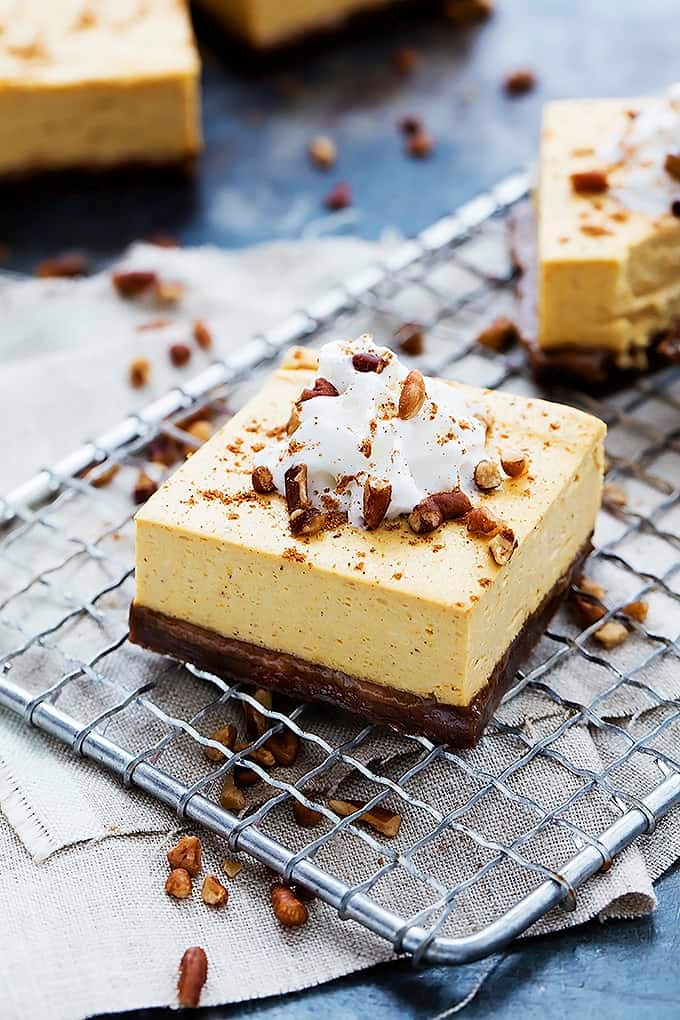 a pumpkin cheesecake bar with gingersnap crust topped with cream and nuts on a cooling rack with. more bars on the side and in the background.