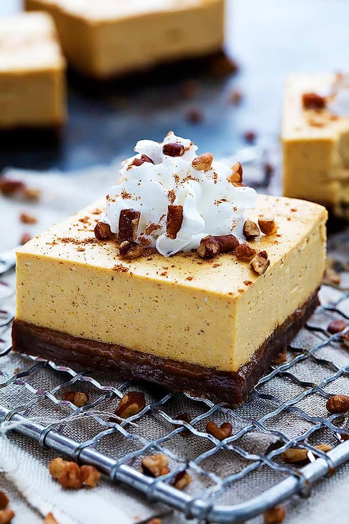 a pumpkin cheesecake bar with gingersnap crust topped with cream and nuts on a cooling rack with more bars in the background.