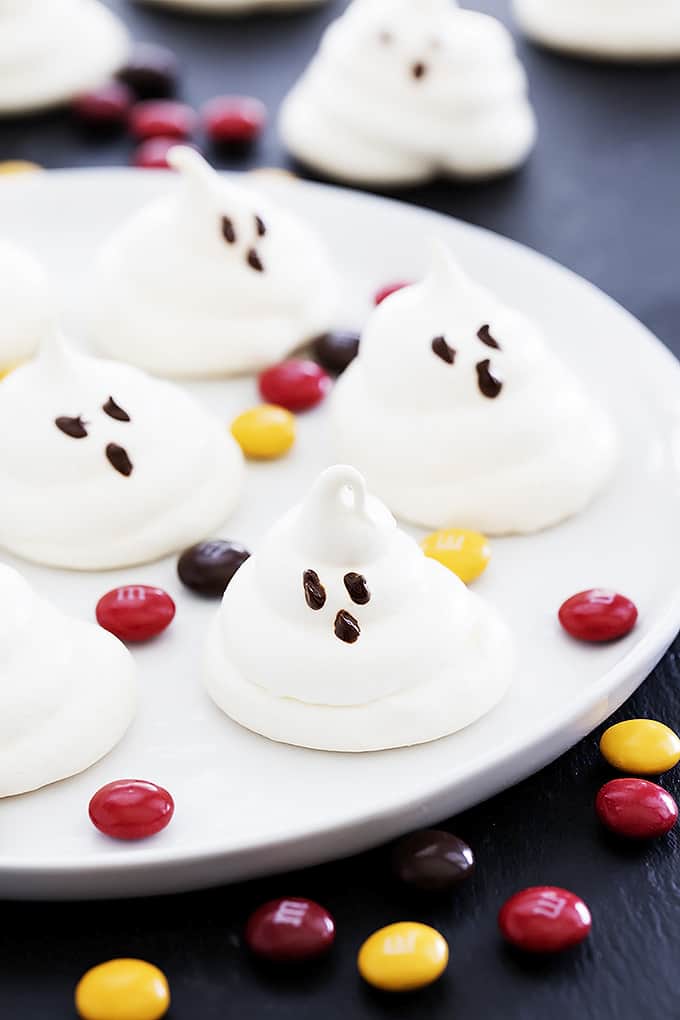 almond meringue ghost cookies on a plate surrounded by m&ms with more cookies and m&ms in the background.