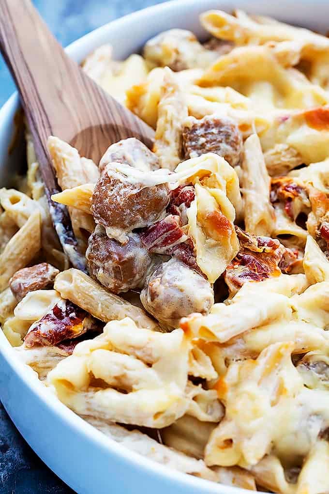 close up of baked cheesy sausage penne with a wooden serving spoon in a bowl.