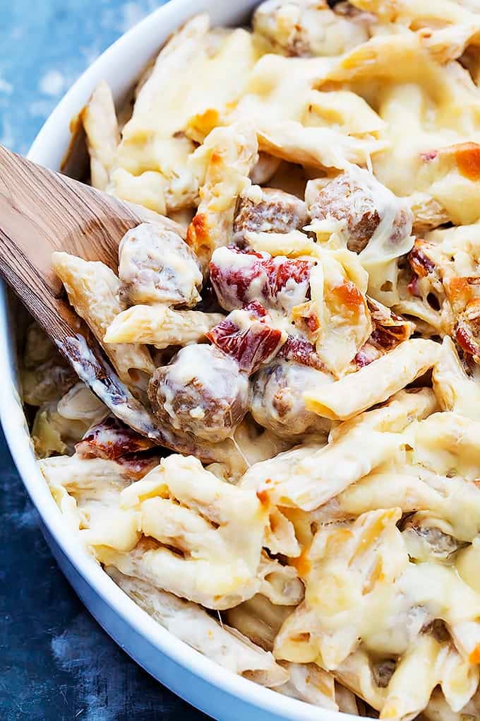 close up of baked cheesy sausage penne and a wooden serving spoon in a bowl.